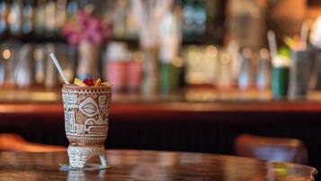 Un ambiente de bar con un vaso decorativo que contiene una bebida y una pajita, una barra y un fondo borroso con botellas y vasos.