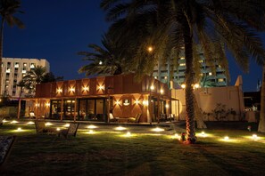 Un edificio moderno con zona de estar al aire libre techada, palmeras y una entrada bien iluminada.