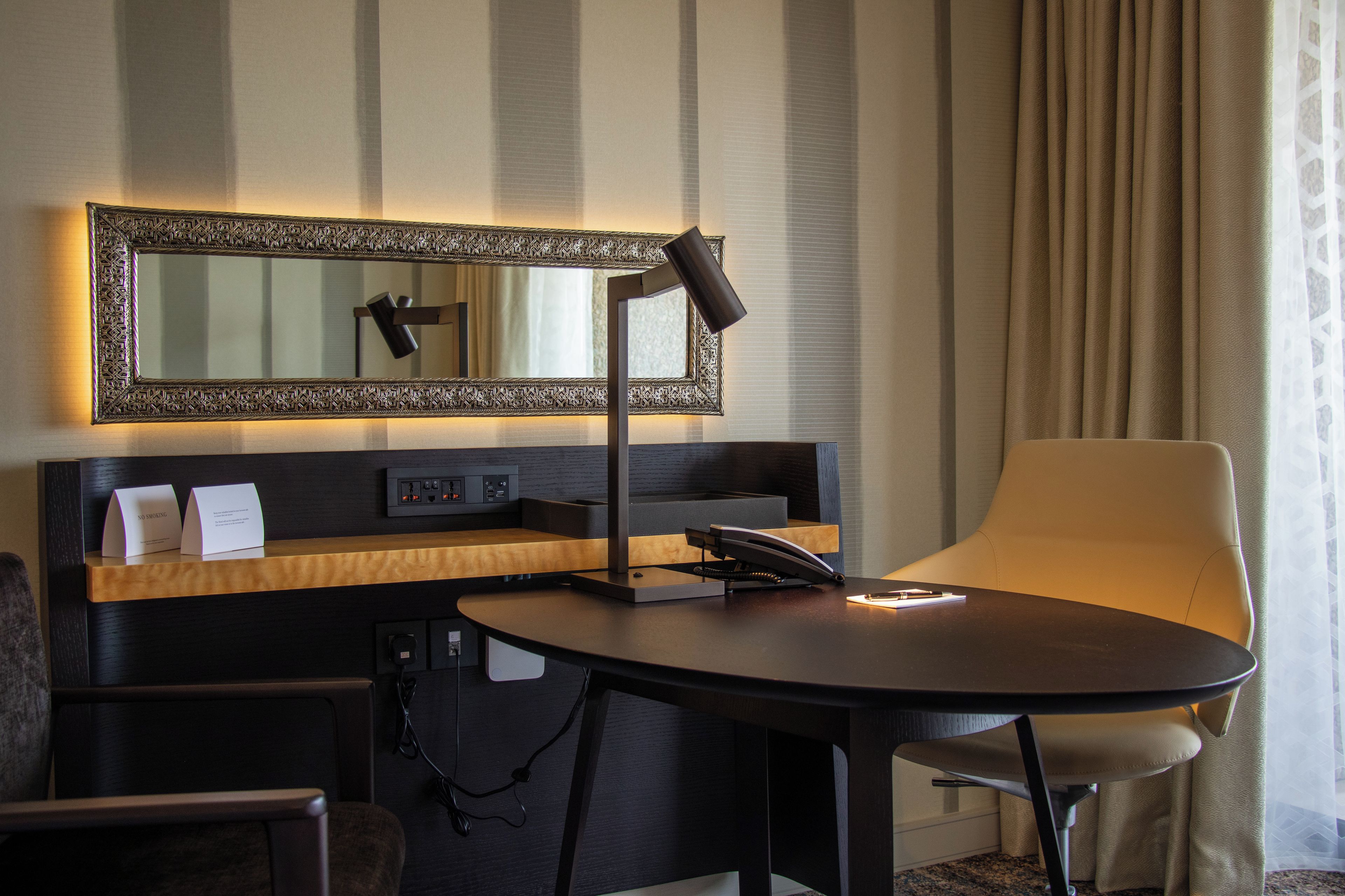A hotel room with a desk, chair, a large mirror, and a telephone.
