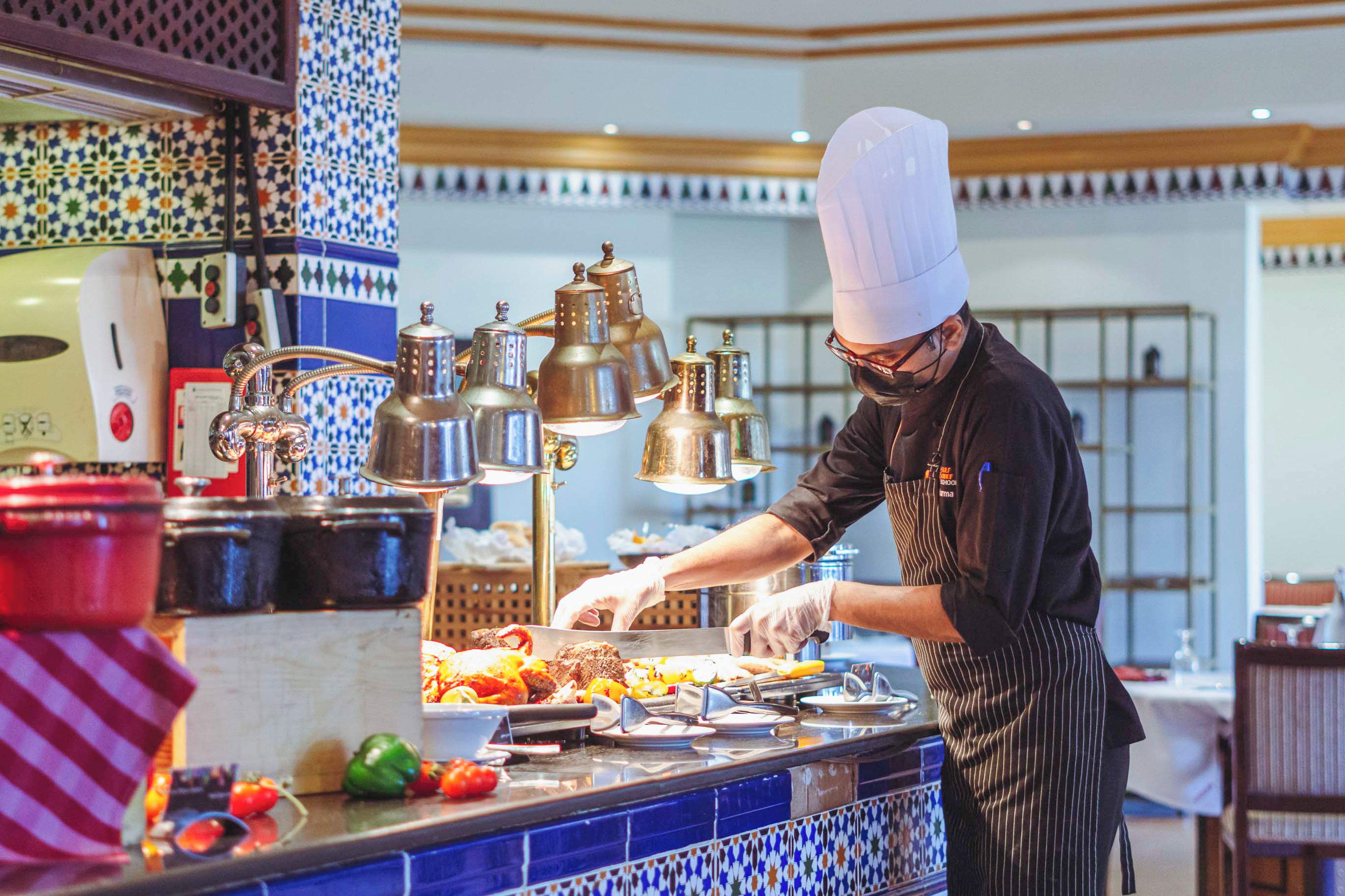 A chef preparing food in a kitchen with blue and white patterned tiles, stainless steel appliances, and a variety of fresh vegetables.