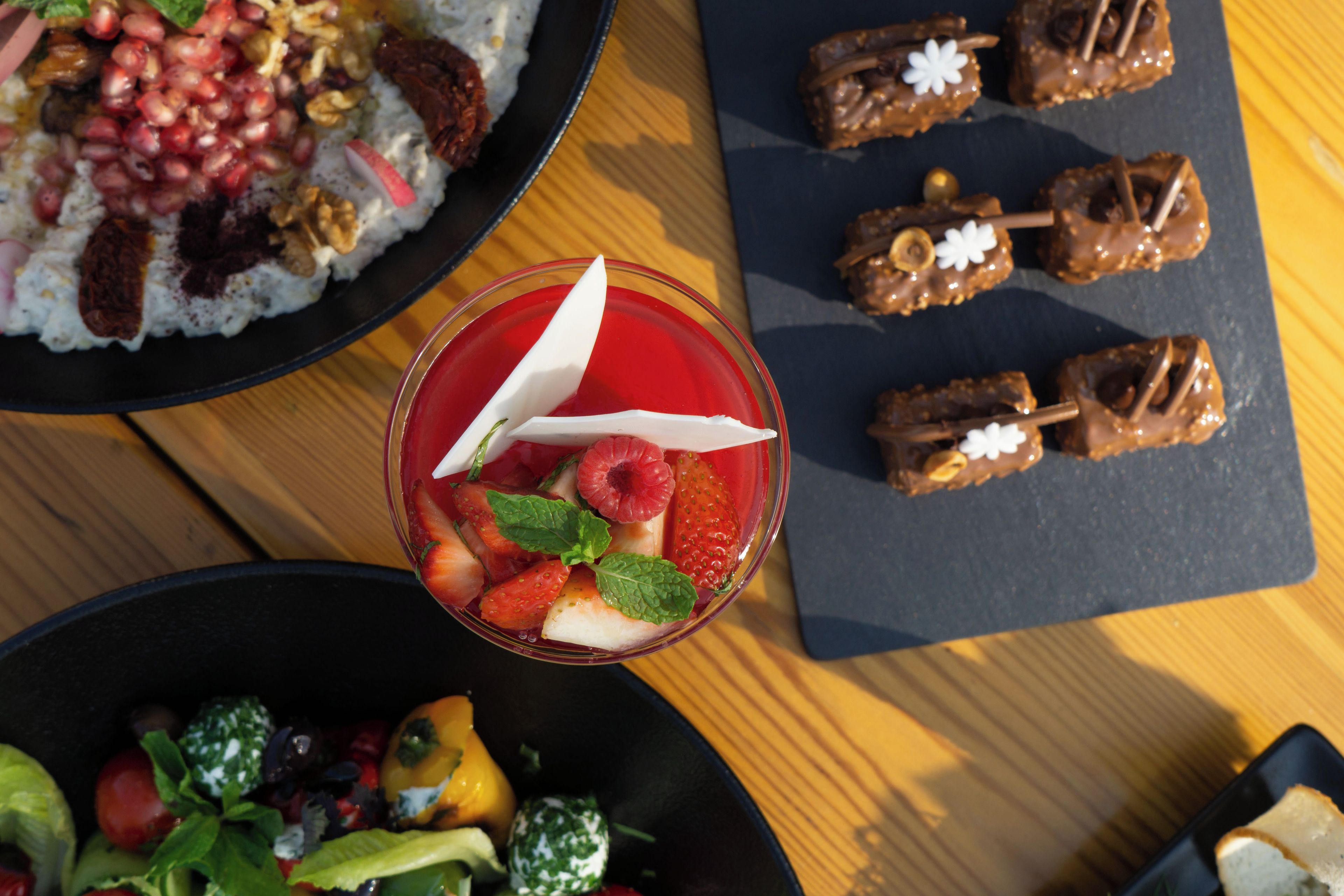Variedad de alimentos sobre una mesa, incluyendo un bol de ensalada de frutas, un plato de arroz con nueces y semillas de granada, y dulces cubiertos de chocolate.