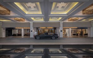 Una entrada de hotel moderna con un coche negro estacionado adelante, amplias ventanas y un vestíbulo espacioso.