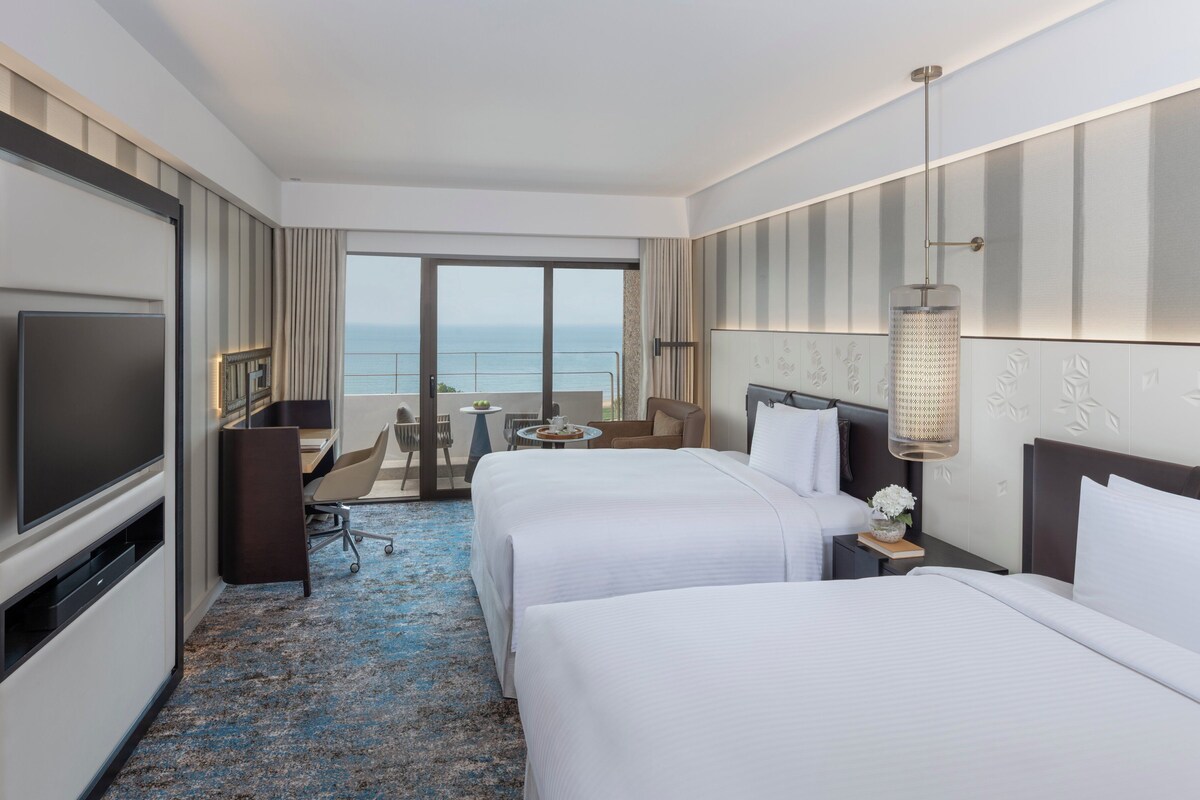 Habitación de hotel con dos camas, televisor, escritorio y balcón con vista al mar.