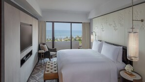A hotel room with a large bed, a TV, a chair, a small table, and a view of the ocean.
