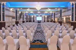 Un salón de conferencias con filas de sillas blancas, piso alfombrado y una gran pantalla al frente.