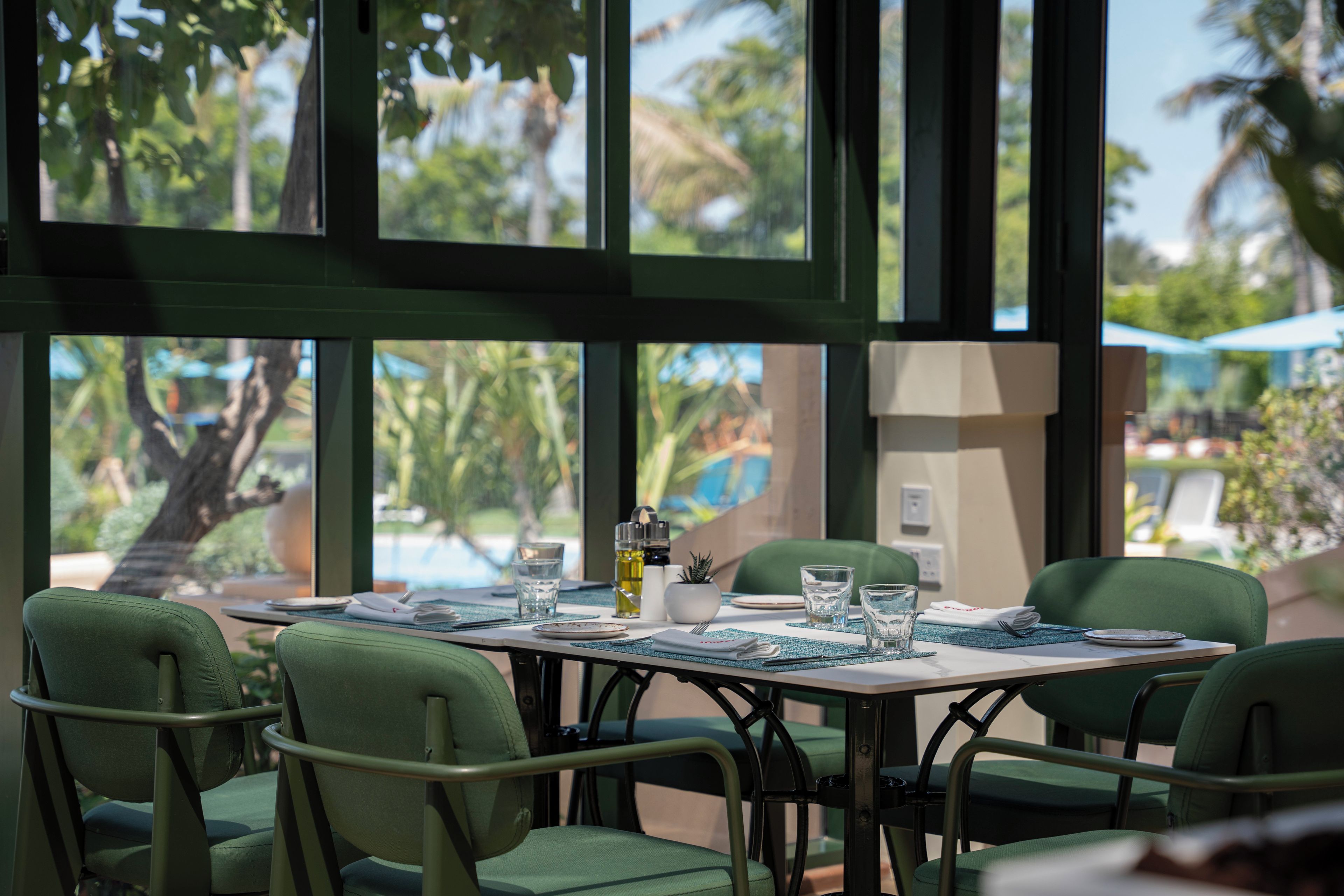 Comedor con una mesa dispuesta para cuatro personas, rodeado de sillas verdes, con vista a palmeras y una piscina exterior.