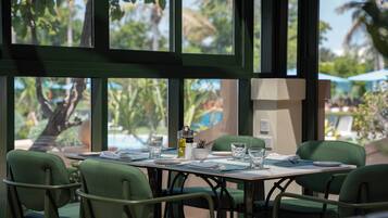 Comedor con una mesa dispuesta para cuatro personas, rodeado de sillas verdes, con vista a palmeras y una piscina exterior.