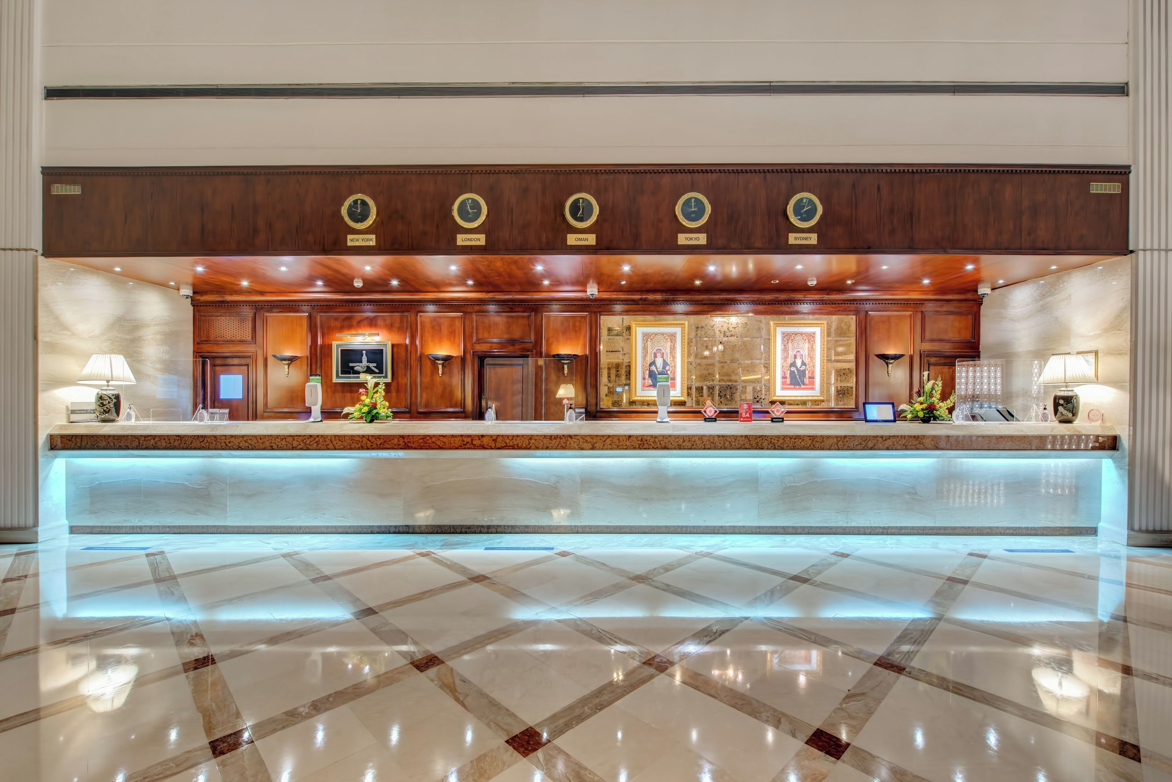 Ein Hotel-Empfangsbereich mit Marmorboden, einer Holztresen, Wanduhren, die verschiedene Zeitzonen anzeigen, und gerahmten Kunstwerken.