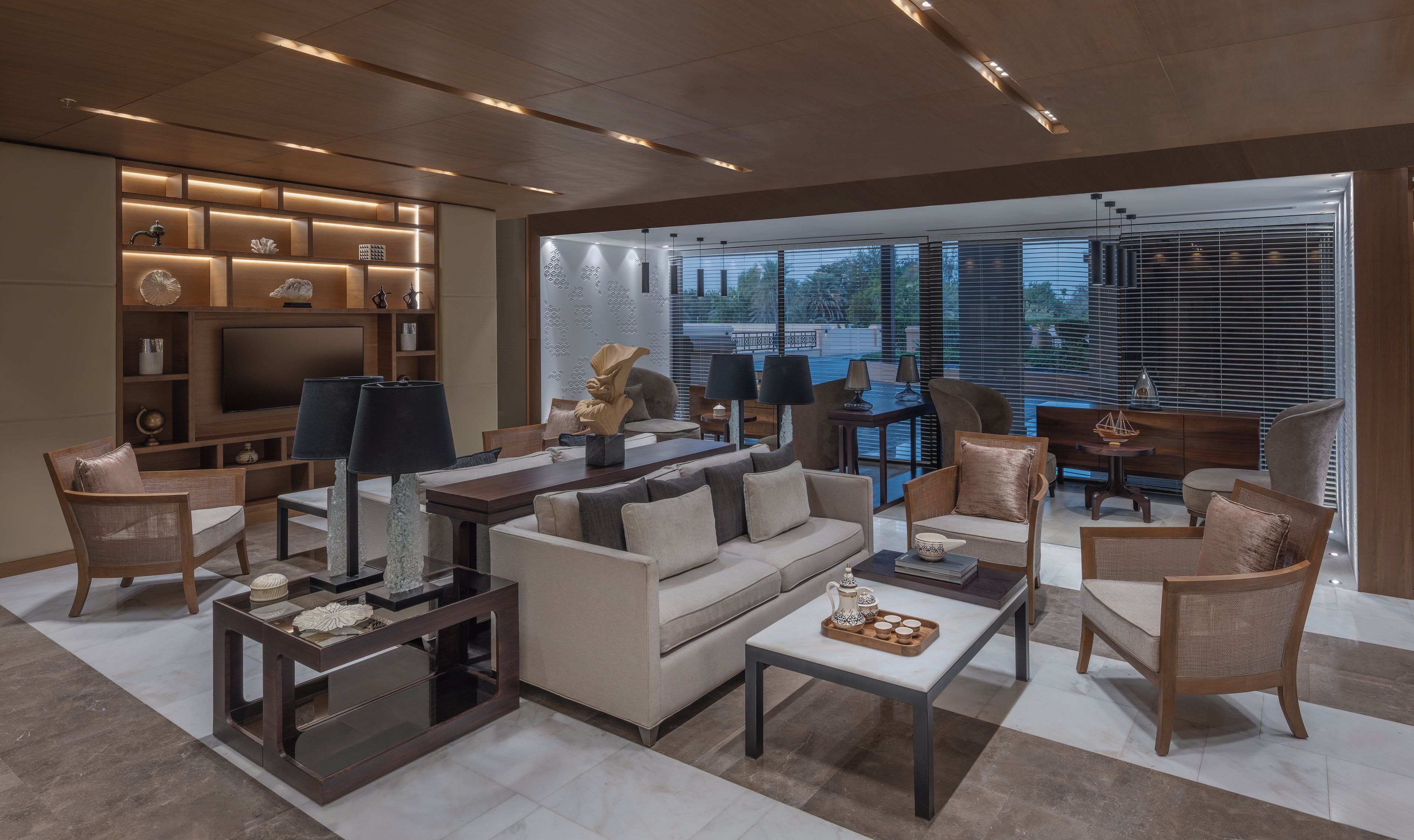 Una sala de estar moderna con un sofá, sillones, una mesa de centro y un televisor. Se observa una estantería con objetos decorativos y un ventanal con persianas.