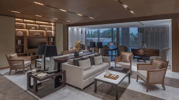 Una sala de estar moderna con un sofá, sillones, una mesa de centro y un televisor. Se observa una estantería con objetos decorativos y un ventanal con persianas.