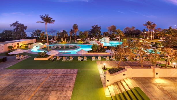A resort pool area with multiple sun loungers and umbrellas, surrounded by palm trees and a well-lit pool. - InterContinental Muscat by IHG (Muscat)