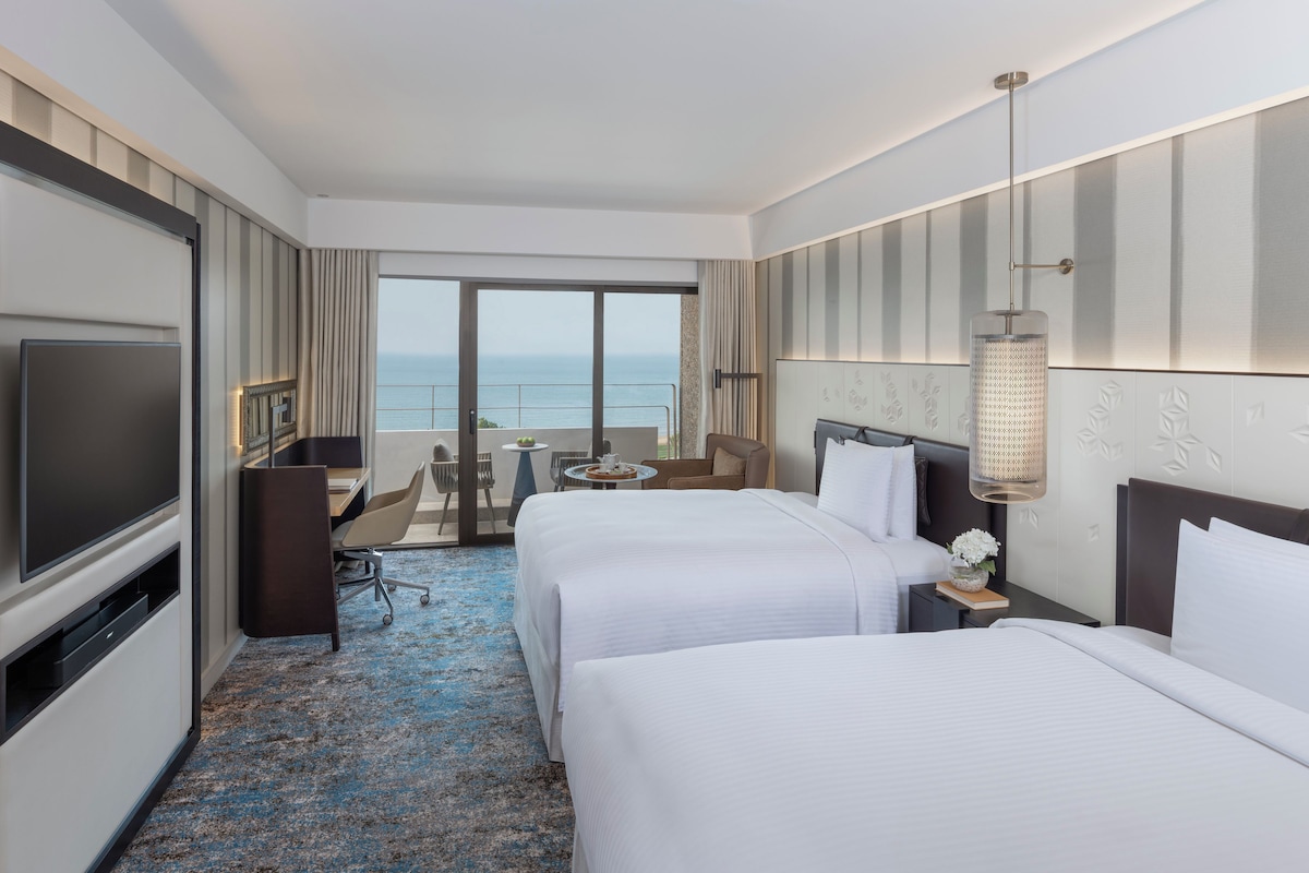 Habitación de hotel con dos camas, televisor, escritorio y balcón con vista al mar.