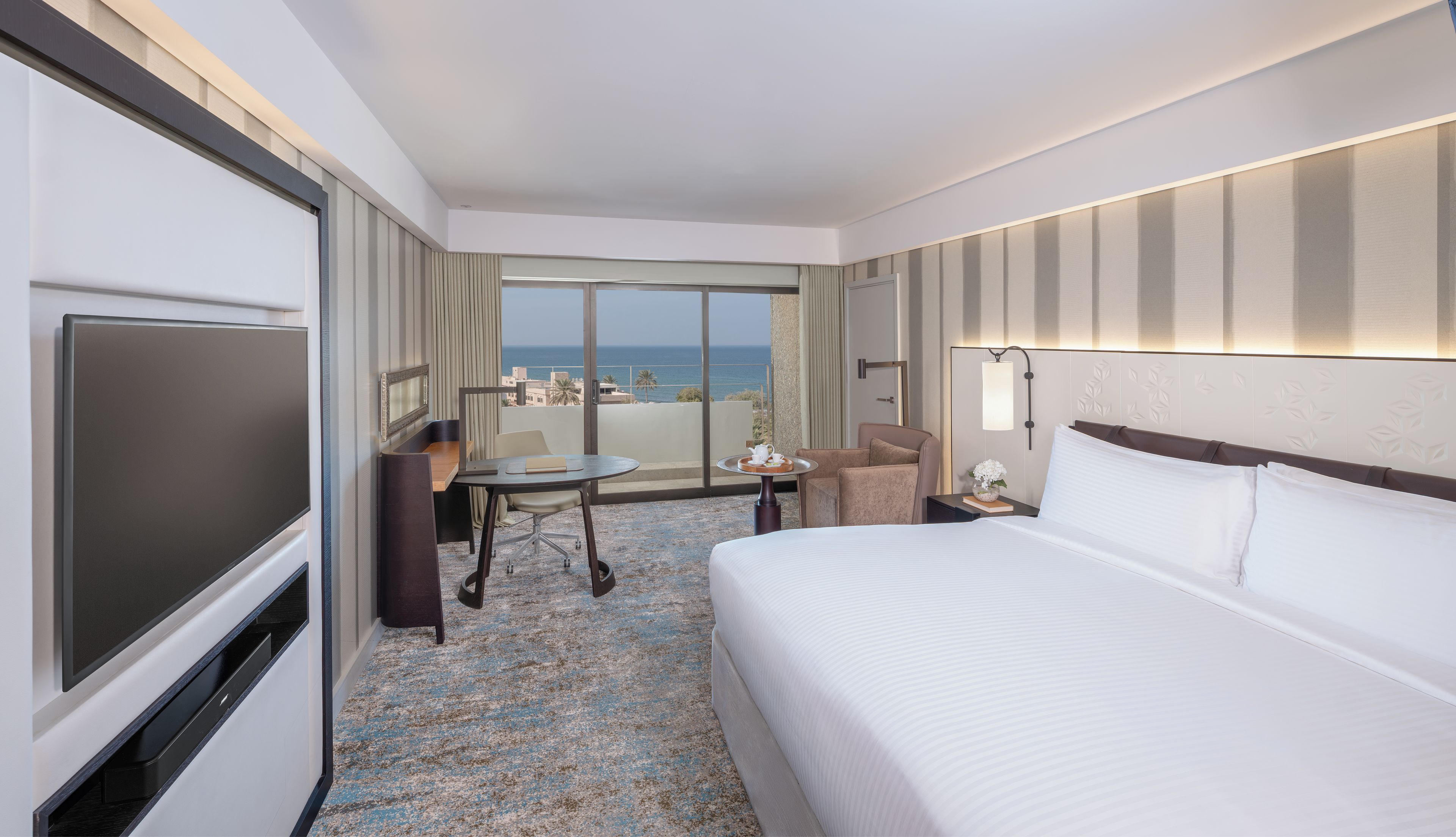 A hotel room with a bed, a desk, a chair, a TV, and a view of the ocean.