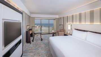 A hotel room with a bed, a desk, a chair, a TV, and a view of the ocean.