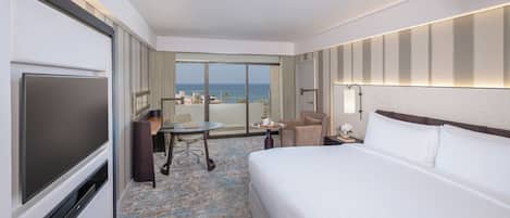 A hotel room with a bed, a desk, a chair, a TV, and a view of the ocean.