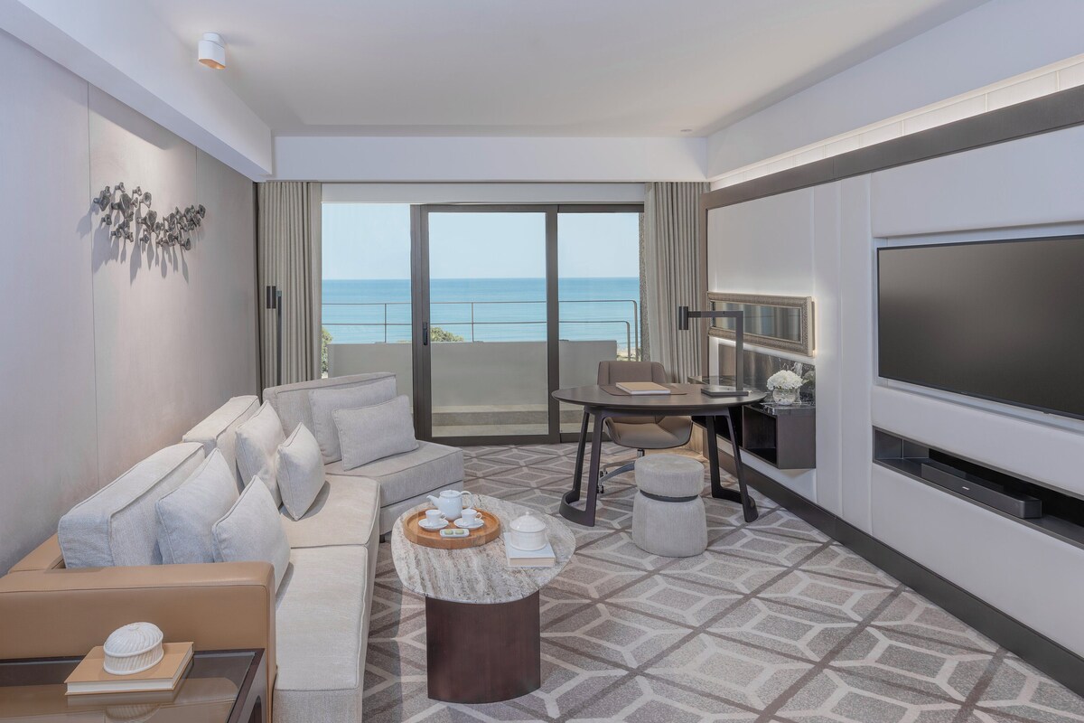 A modern hotel room with a sofa, a dining table, a TV, and a balcony with a view of the sea.
