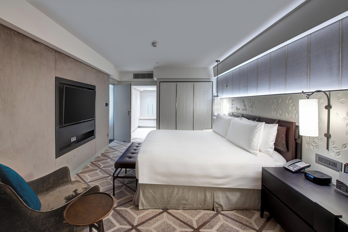 A hotel room with a large bed, a TV mounted on the wall, a bedside table with a lamp, a bench, and a patterned carpet.