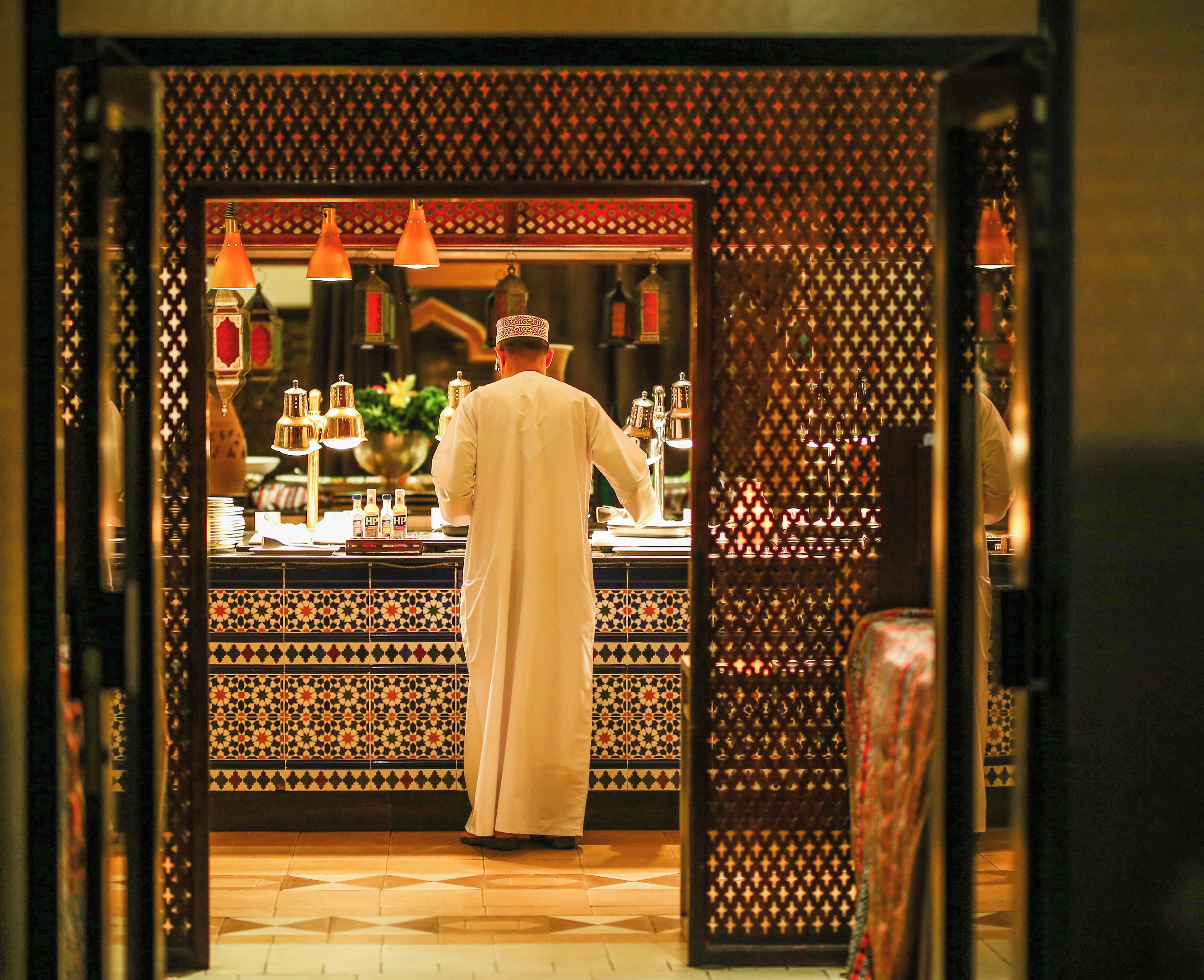 A person in traditional attire working behind a counter with a patterned backdrop and hanging lights.