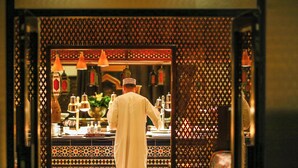 A person in traditional attire working behind a counter with a patterned backdrop and hanging lights.