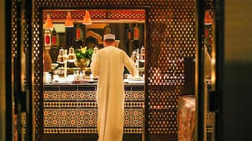 A person in traditional attire working behind a counter with a patterned backdrop and hanging lights.