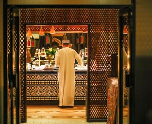 A person in traditional attire working behind a counter with a patterned backdrop and hanging lights. - InterContinental Muscat by IHG (Muscat)