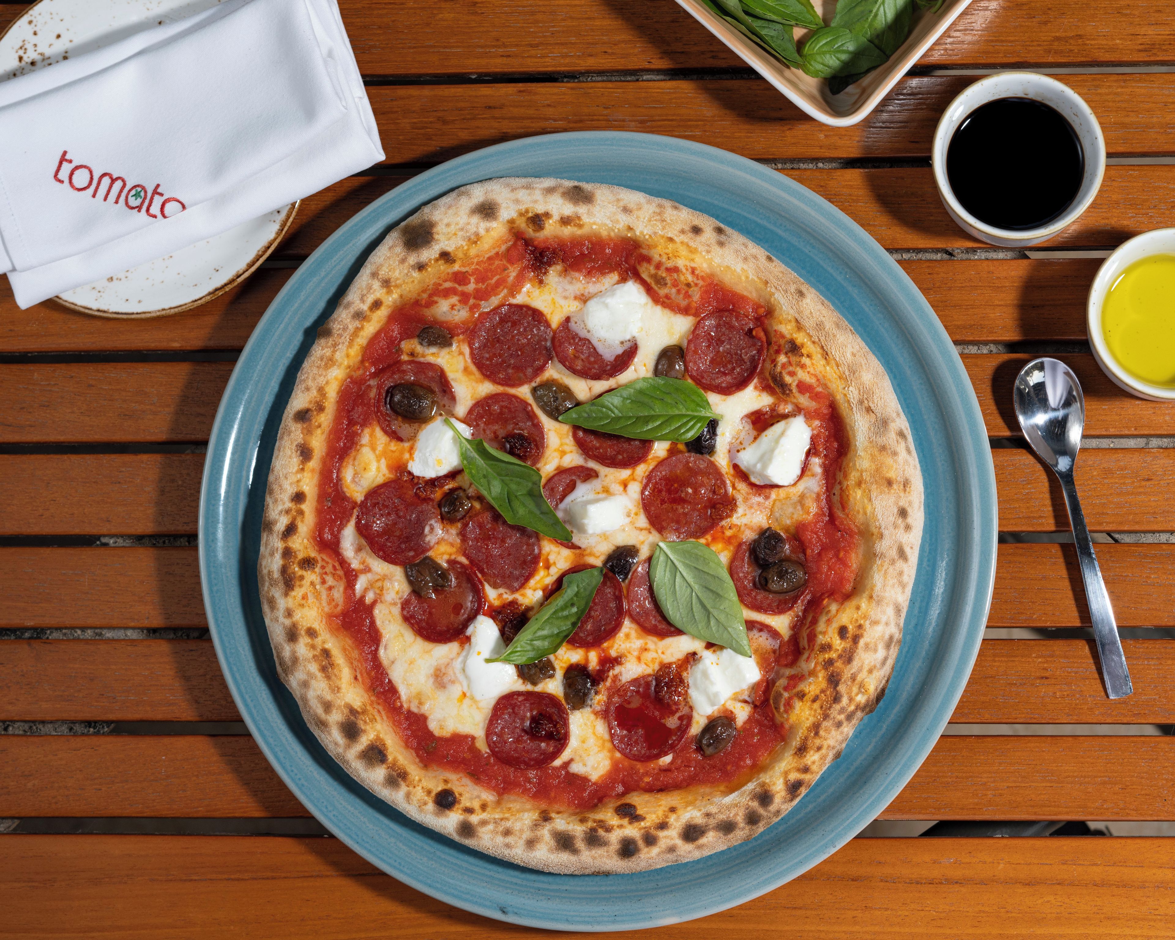 Una pizza en un plato azul, con pepperoni, queso y albahaca, sobre una mesa de madera.