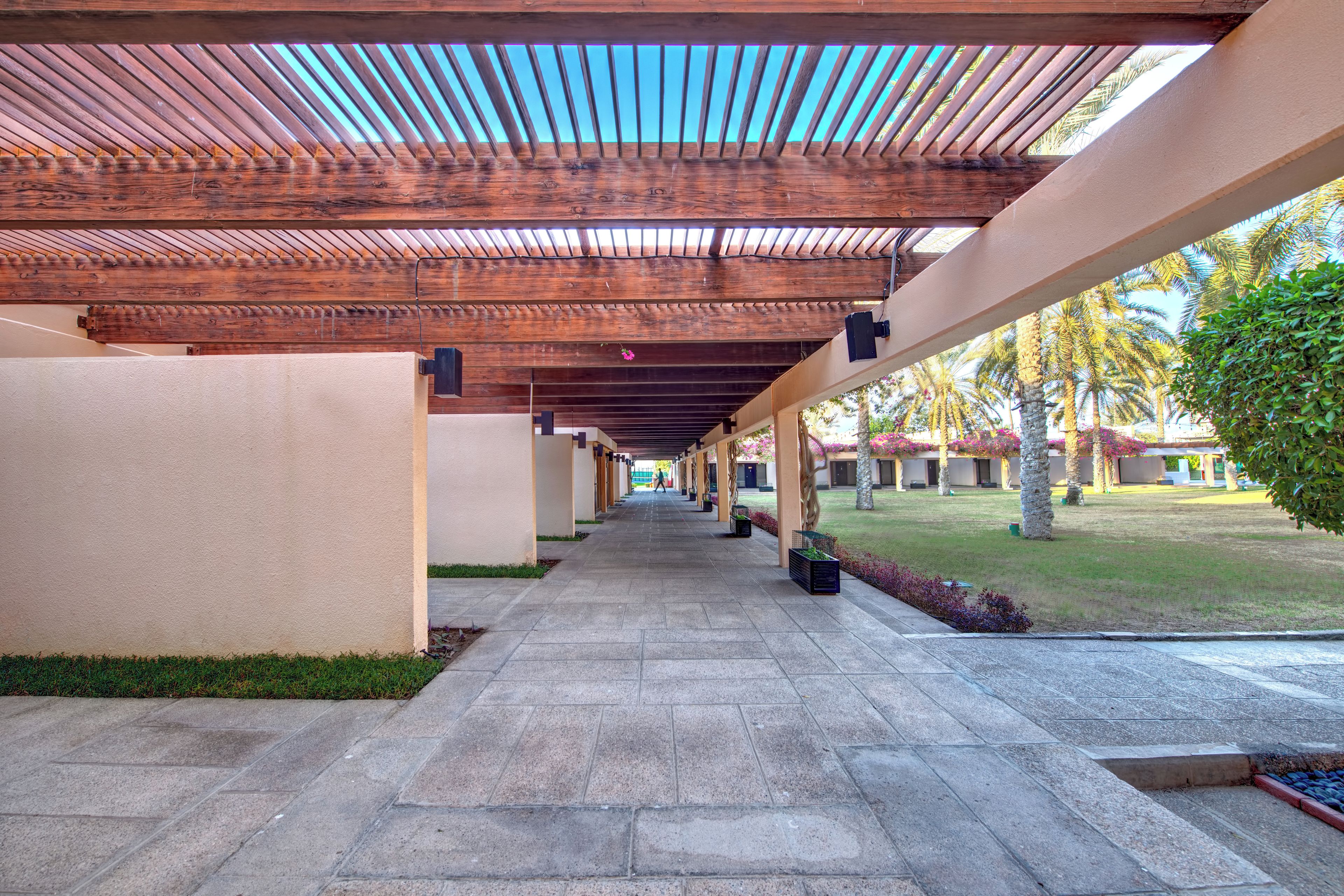 A covered walkway with a tiled floor, wooden beams, and a view of a landscaped area with palm trees.