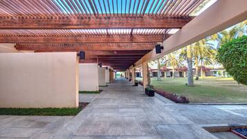A covered walkway with a tiled floor, wooden beams, and a view of a landscaped area with palm trees.