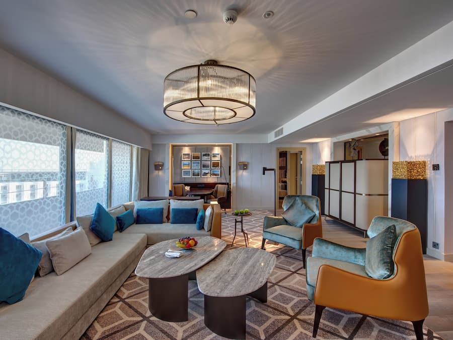 Una habitación de hotel moderna con un sofá grande, dos sillones, una mesa de centro y un comedor con una cocina al fondo.
