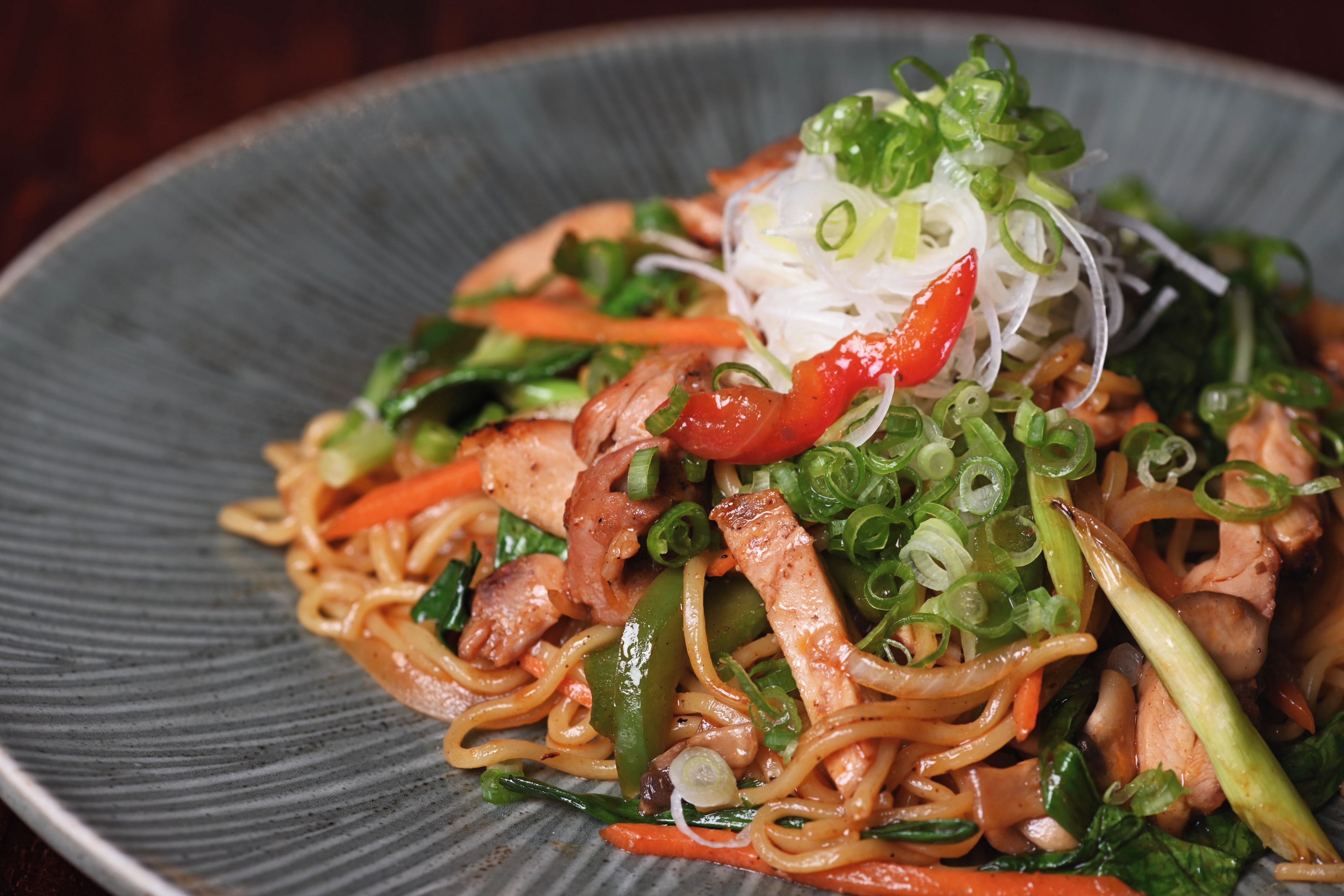 A plate of stir-fried noodles with vegetables and meat, garnished with sliced green onions and red chili.