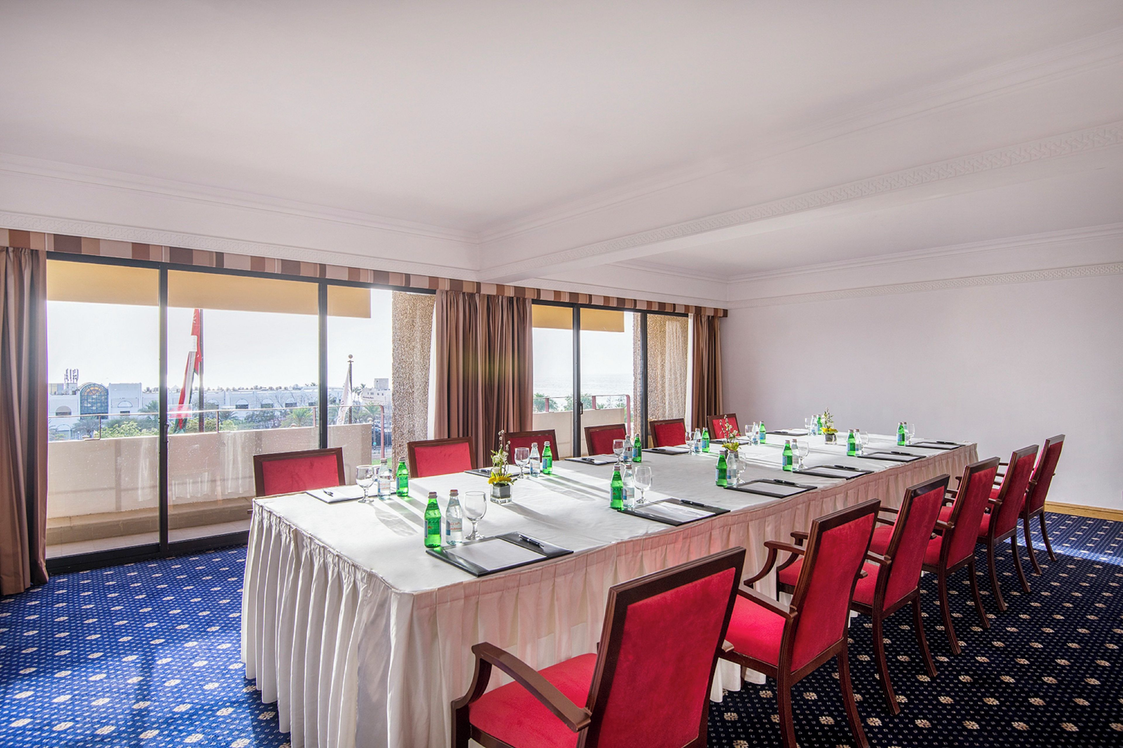 Un salón de conferencias con una larga mesa, sillas rojas y vistas a un paisaje urbano a través de amplios ventanales.