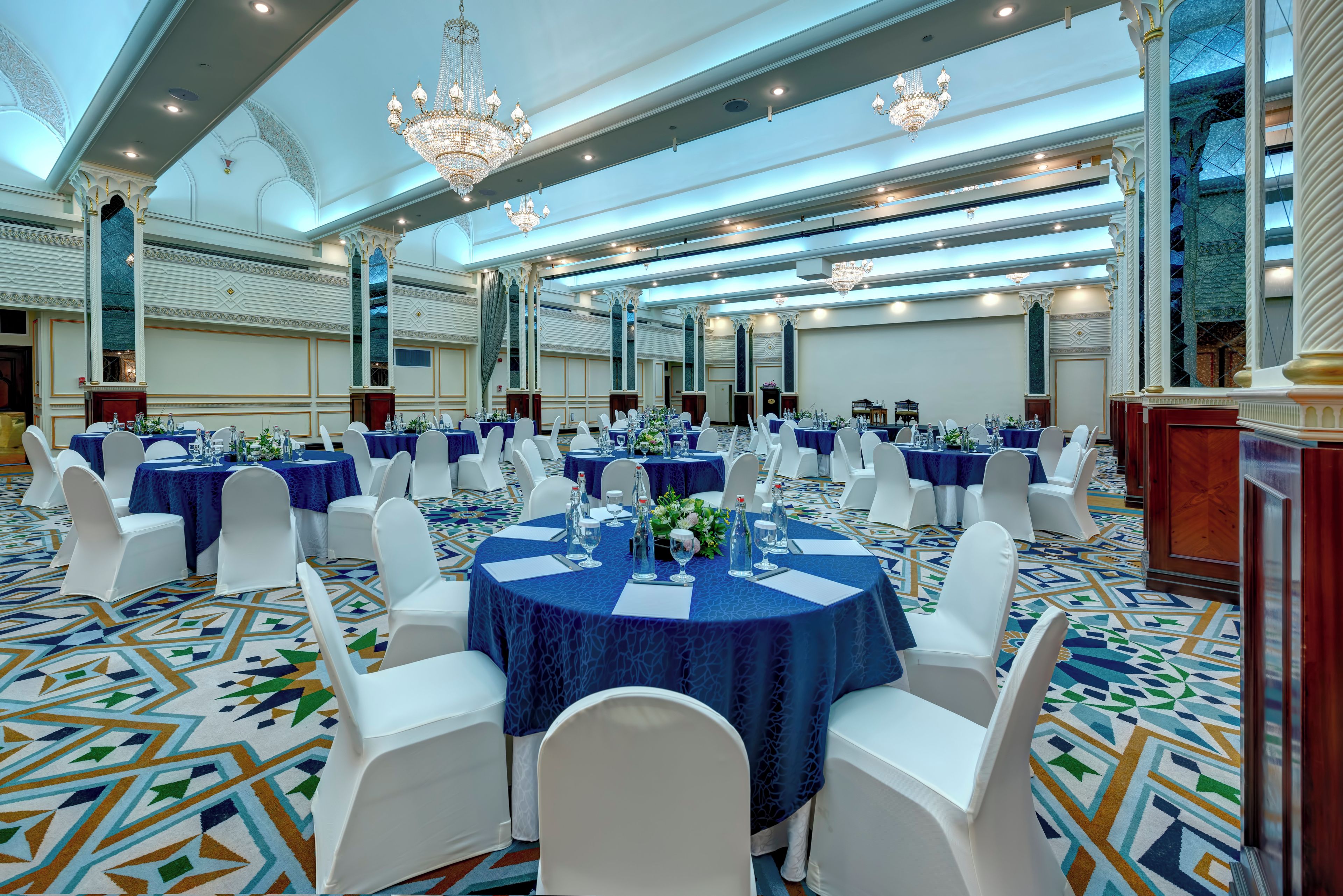 Un salón de banquetes con mesas redondas, sillas blancas y candelabros.