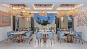 A modern dining area with multiple tables and chairs, large hanging lamps, and a view of palm trees through large windows.