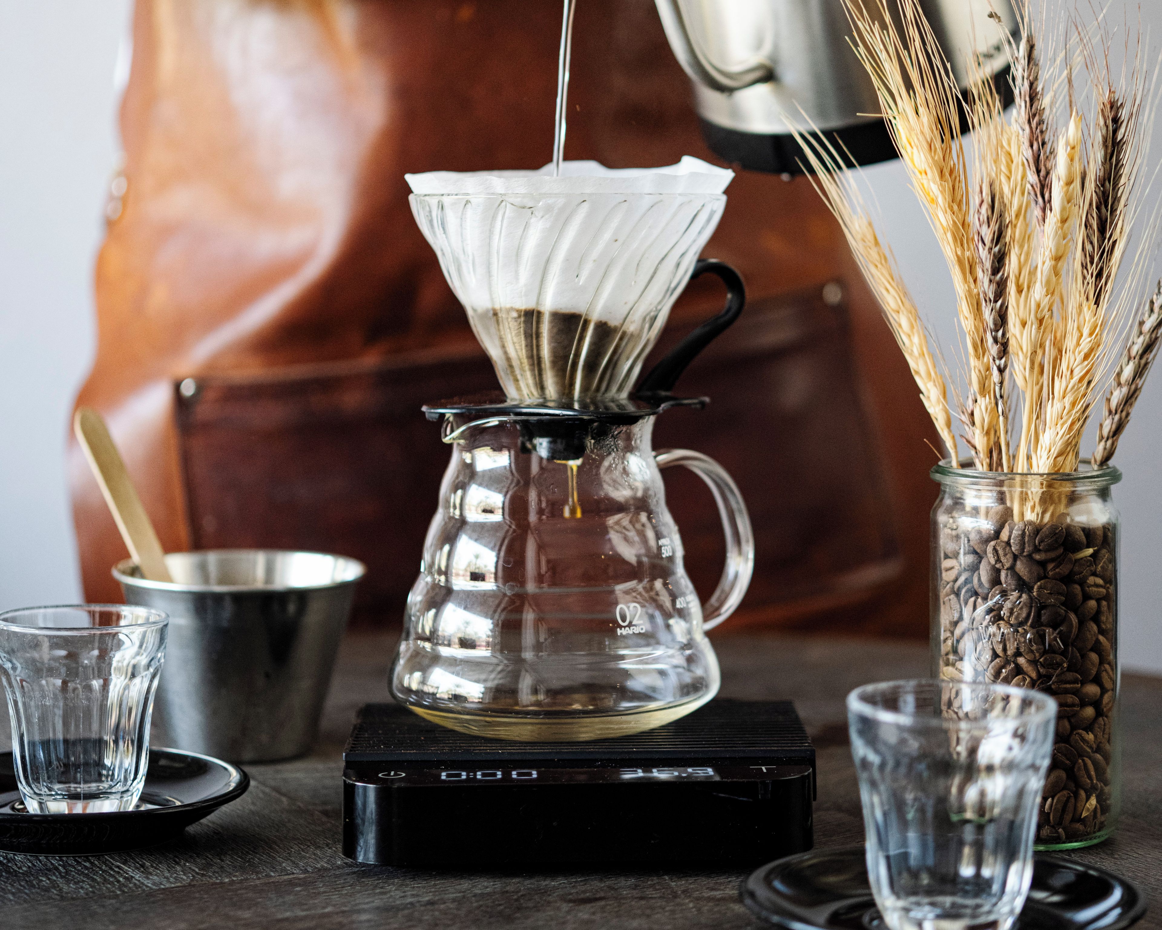 Una cafetera con jarra de vidrio transparente, un tarro de vidrio con granos de café y una cuchara en un recipiente metálico.