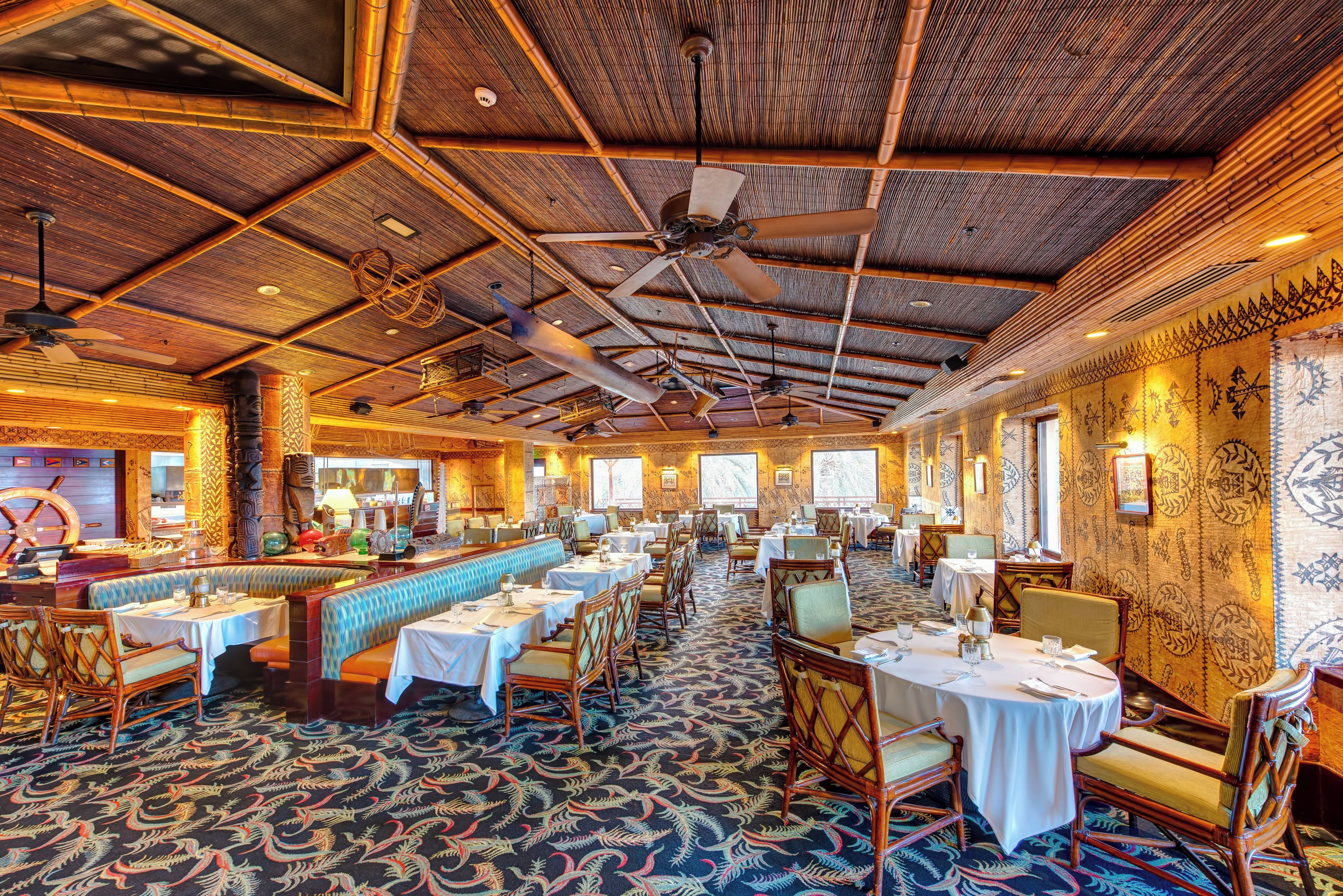A restaurant with high ceilings, bamboo beams, and a patterned carpet. Tables are set with white tablecloths and chairs.