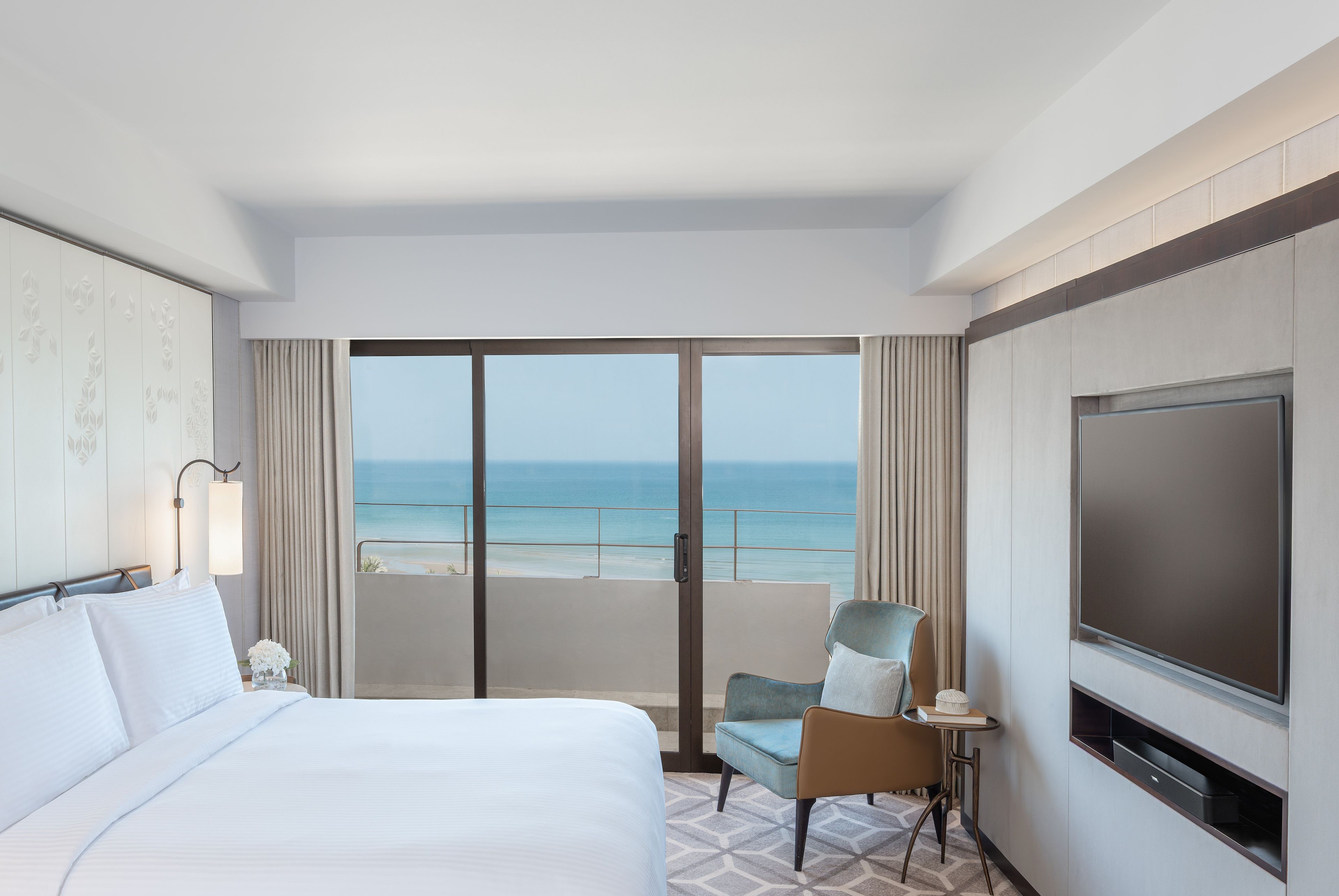 A hotel room with a bed, a chair, a TV, and a balcony with a view of the ocean.