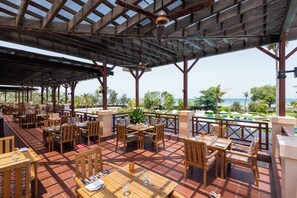 An outdoor restaurant with wooden tables and chairs, a pool in the background, and a view of the ocean. - InterContinental Muscat by IHG (Muscat)