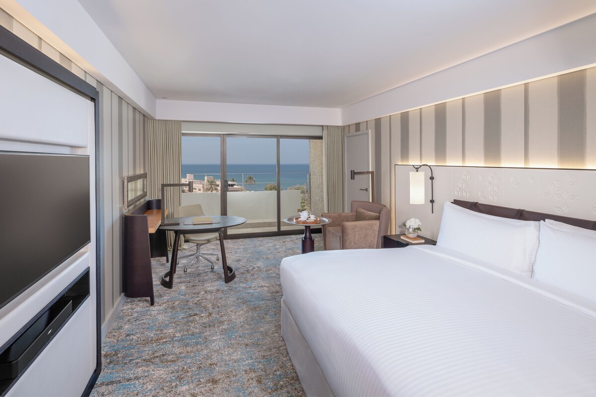 Habitación de hotel con una cama grande, un escritorio con silla, televisor y vista al océano.