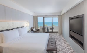 A hotel room with a large bed, a TV, a bench, a small table with chairs, and a balcony with a view of the ocean. - InterContinental Muscat by IHG (Muscat)