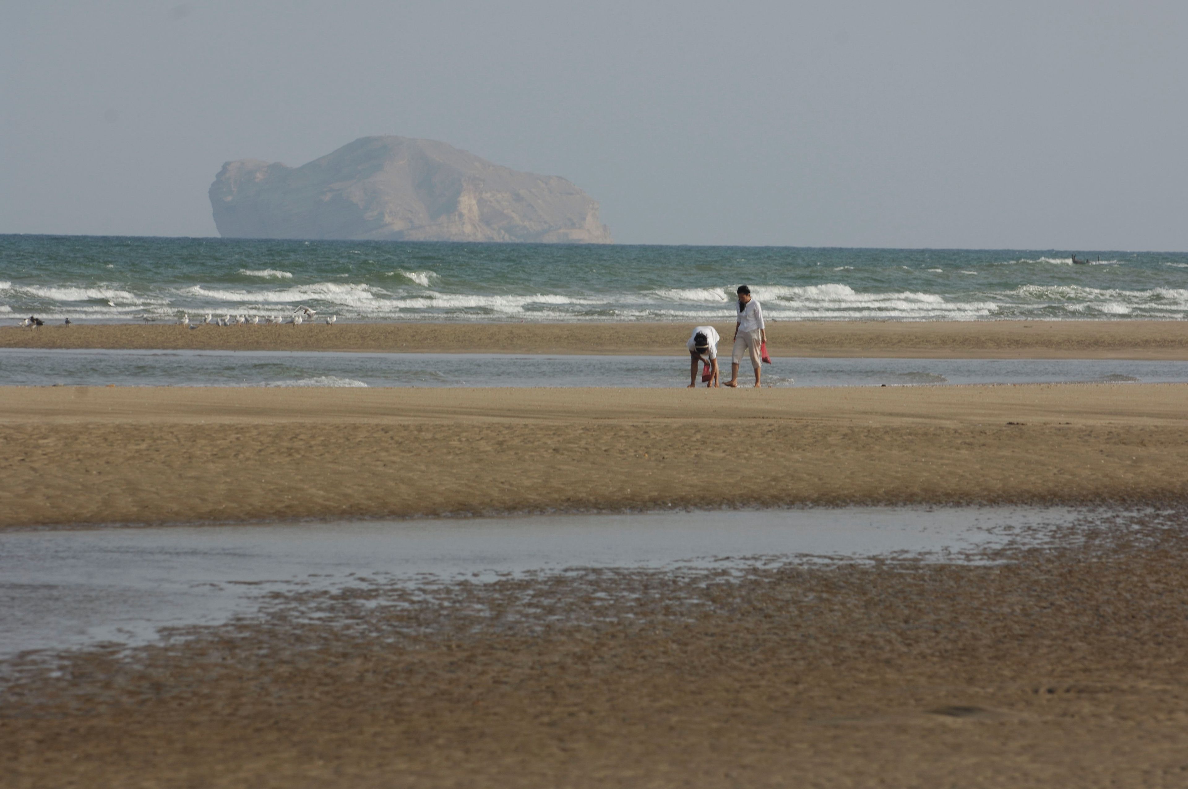 Ein Strand mit einer großen Felsformation im Hintergrund, einem Sandufer und einigen Personen.