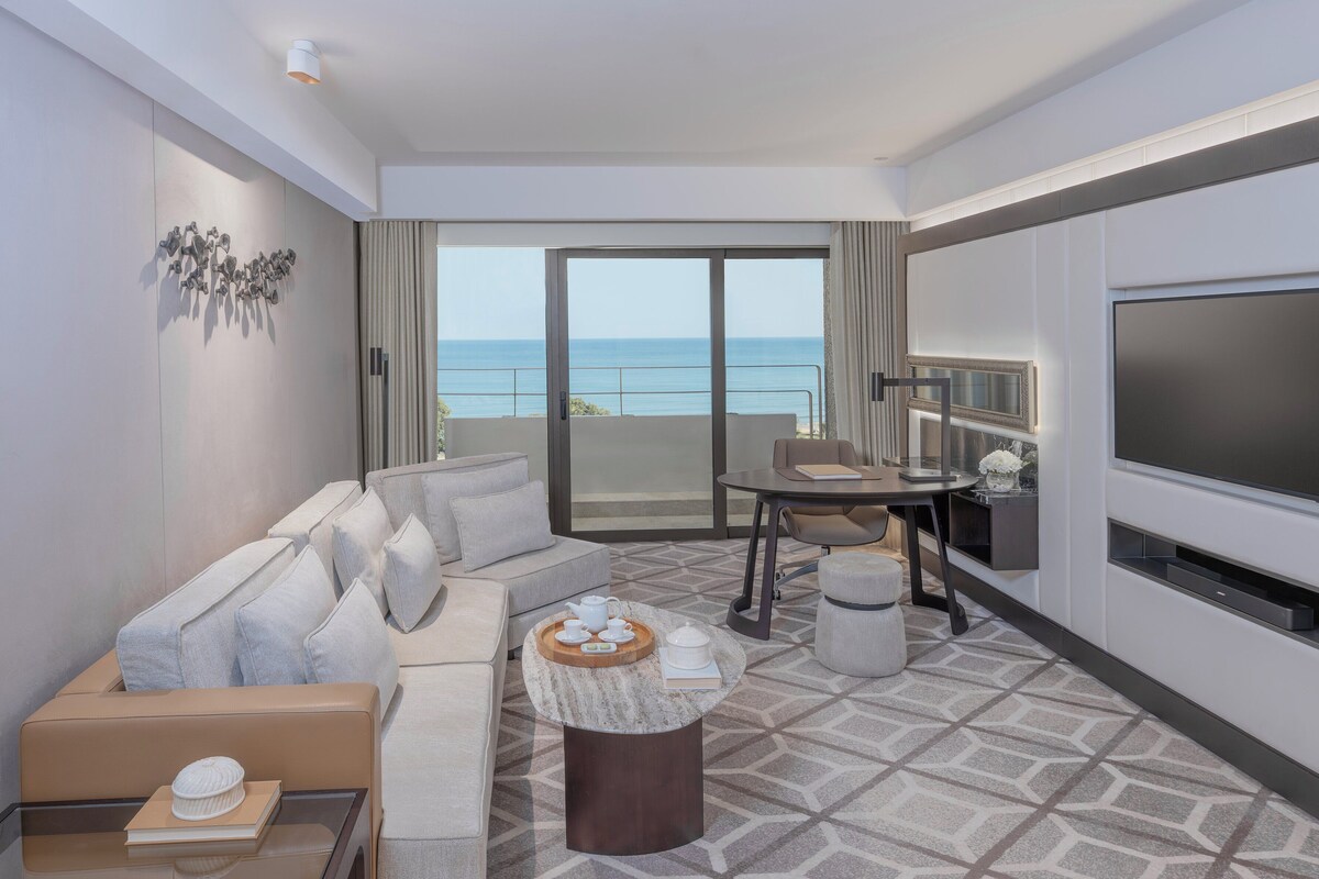 Una habitación de hotel moderna con sofá, una mesa de comedor, televisión y balcón con vistas al mar.