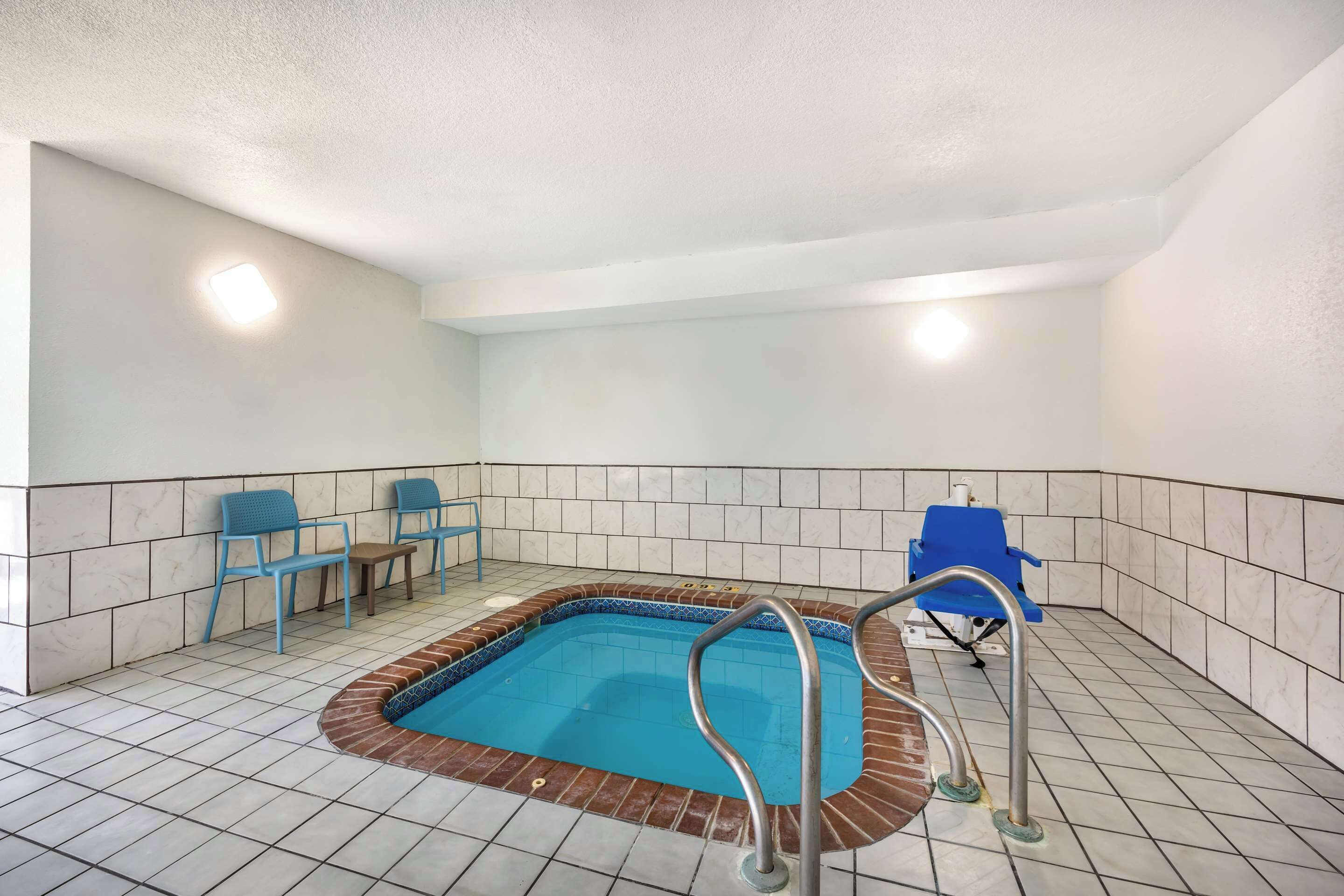 A swimming pool with a tiled floor, a brick border, and a metal ladder. There are blue chairs and a blue chair in the background.