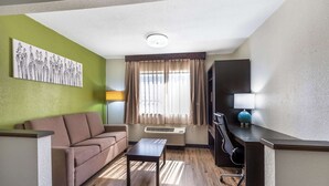 A hotel room with a brown sofa, a small table, a desk with a lamp, and a large window with curtains.