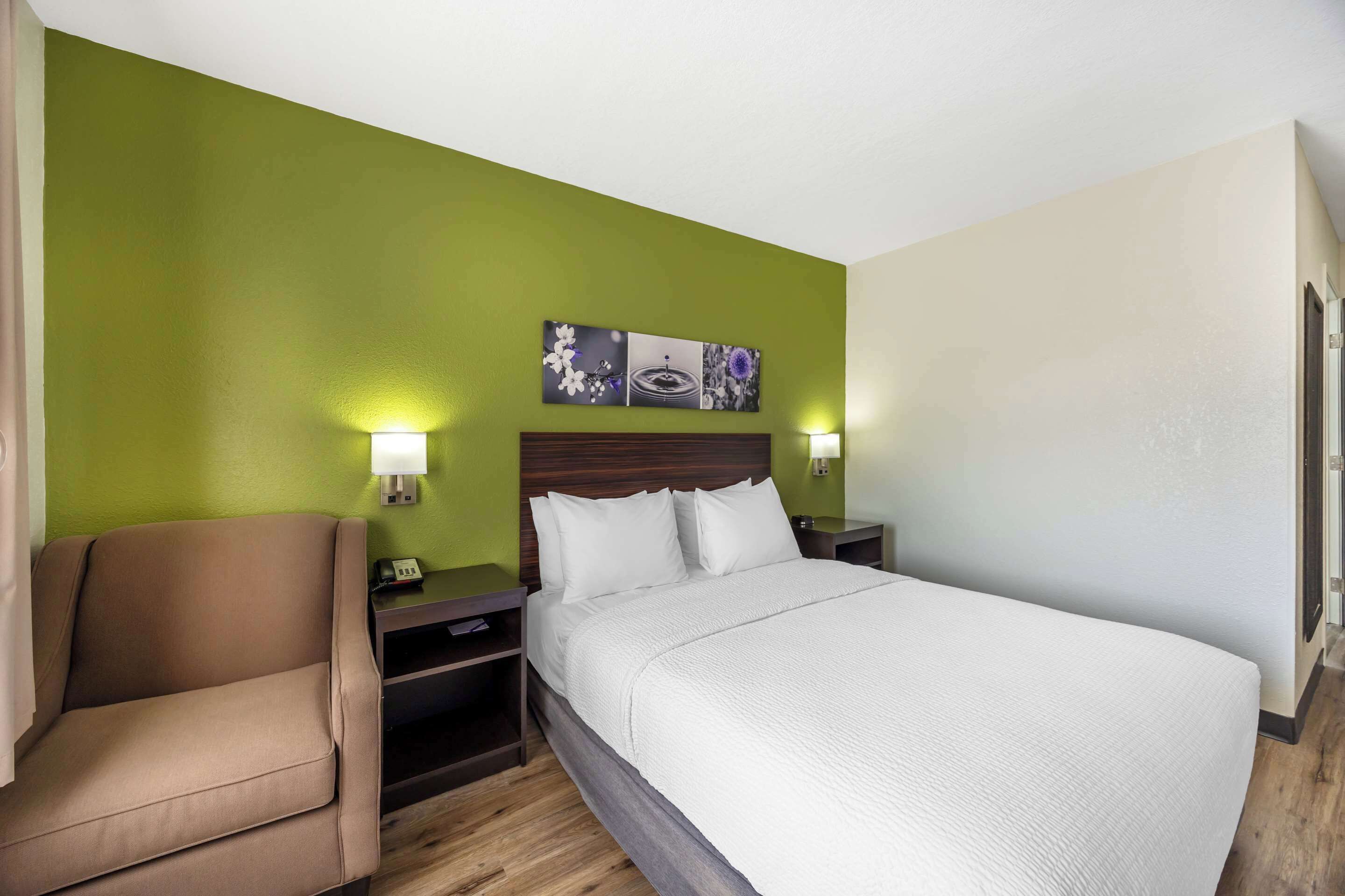 Quarto de hotel com uma cama, uma cadeira e uma mesa de cabeceira com abajur.