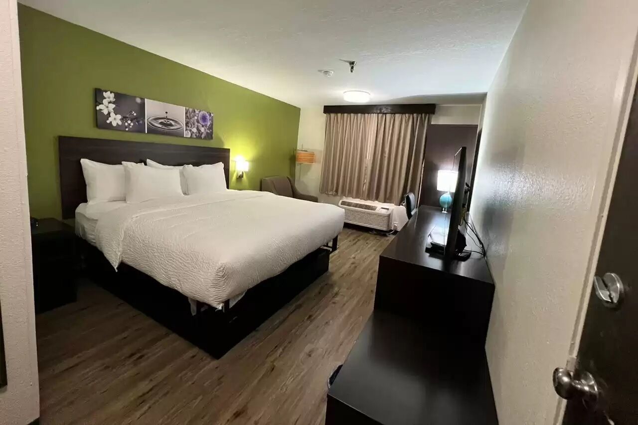 Une chambre d’hôtel avec un lit, un bureau, une chaise, une lampe et une fenêtre avec des rideaux.