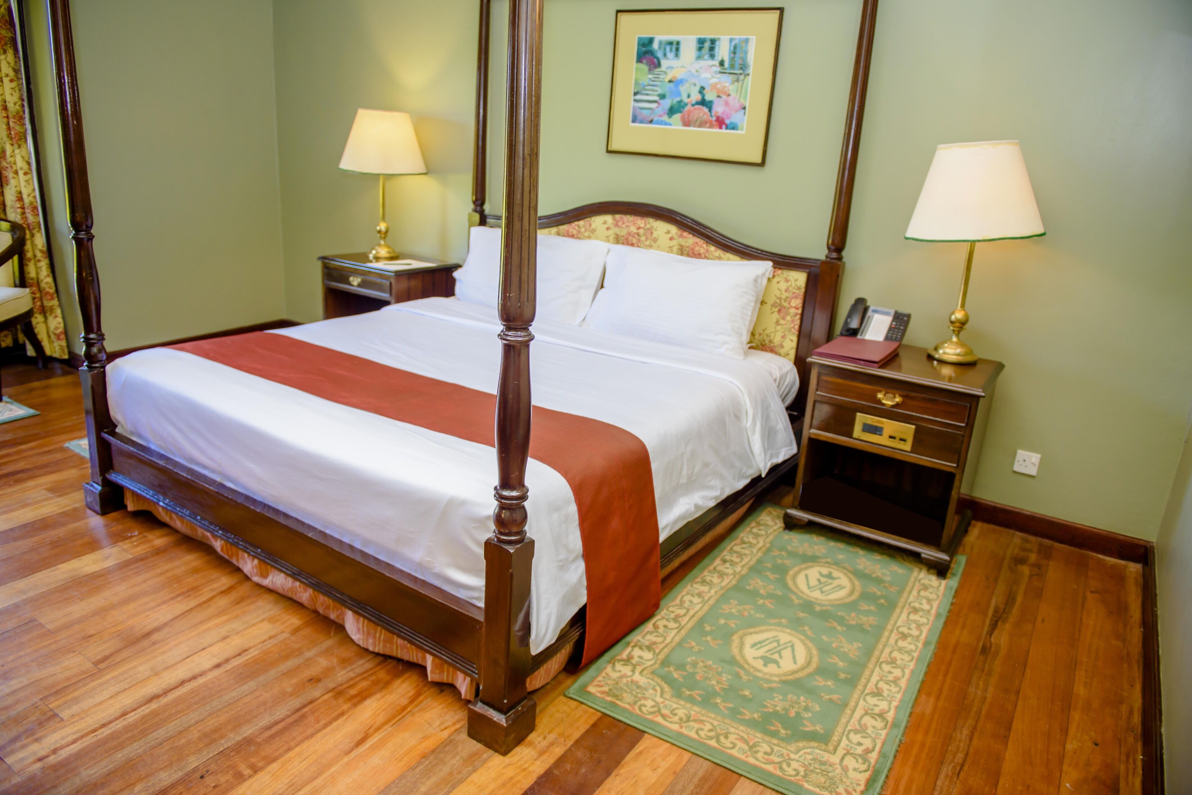 A four-poster bed with a red bedspread, two bedside tables with lamps, a green patterned rug, and a framed picture on the wall.