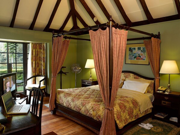 A four-poster bed with floral bedding, a laptop on a chair, and a view of greenery through large windows. - Windsor Golf Hotel & Country Club (Nairobi)