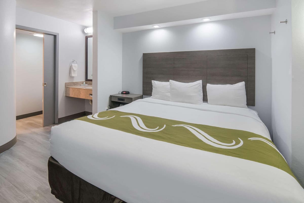 A hotel room with a bed, a nightstand, a mirror, and a bathroom with a sink and towel rack.