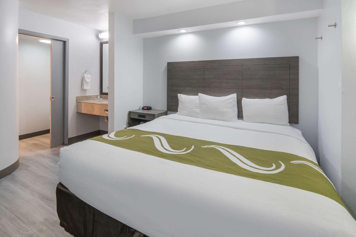 A hotel room with a bed, a nightstand, a mirror, and a bathroom with a sink and towel rack.