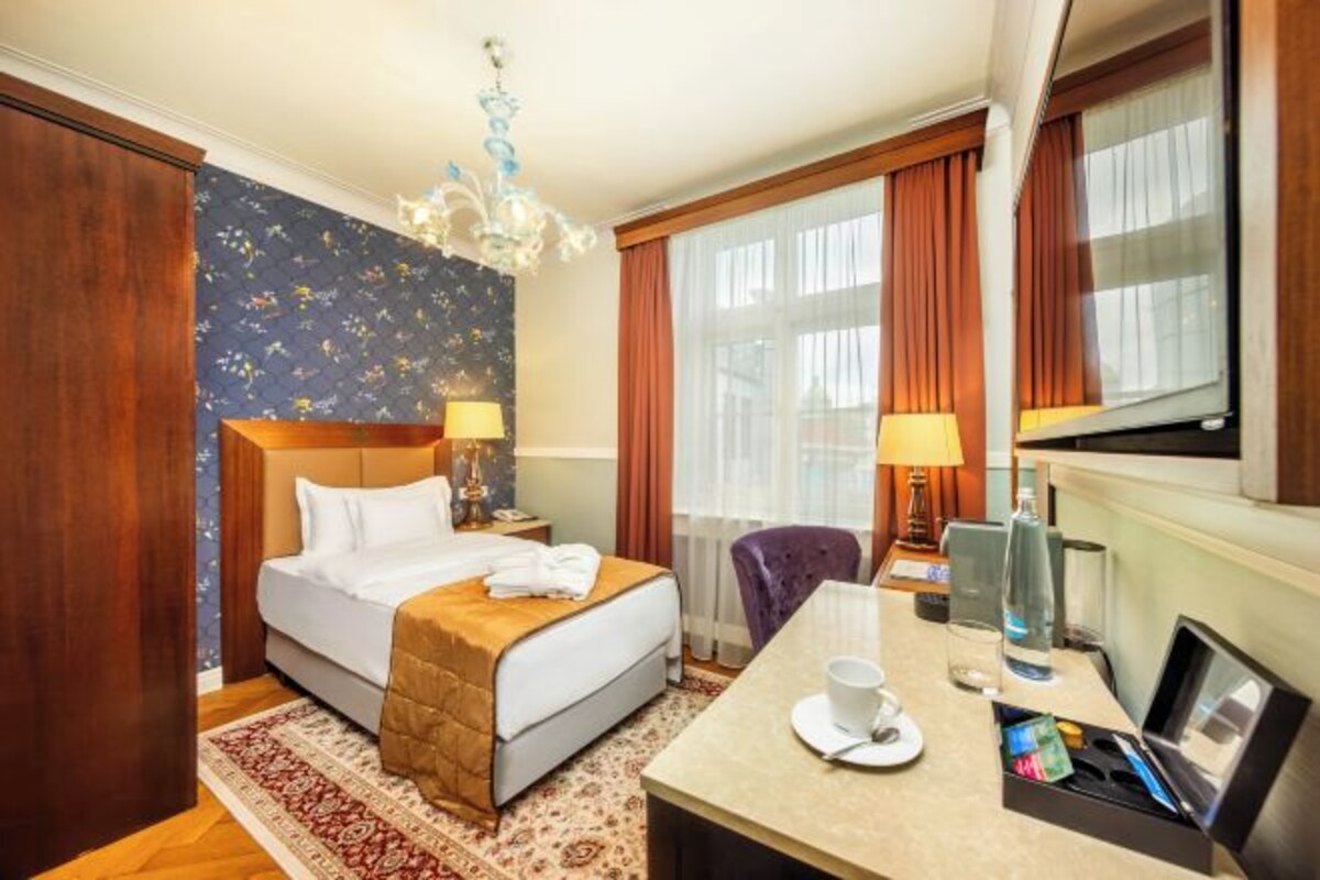 A hotel room with a bed, bedside tables, a desk with a chair, a window with curtains, and a chandelier.
